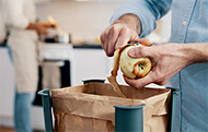 Händer som skalar ett äpple över en matavfallspåse  i ett kök. En person i bakgrunden står vid spisen i förkläde.