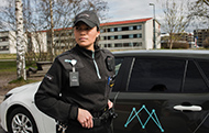 Kvinna i keps och svarta uniformskläder och id-bricka framför svart och vit bil med Avarns turkosa logotyp på