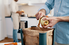 Händer som skalar ett äpple över en matavfallspåse i ett kök. En person i bakgrunden står vid spisen i förkläde.