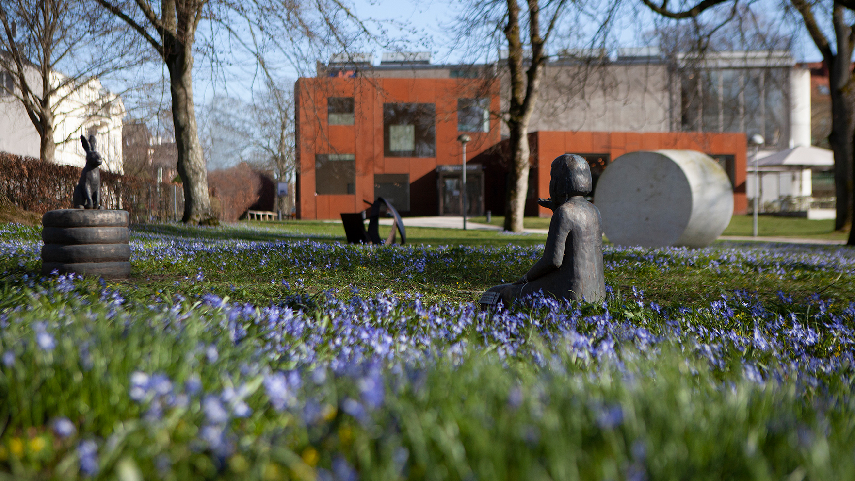 Skulpturer placerade i en park med blå vårblommor och byggnader i bakgrunden.