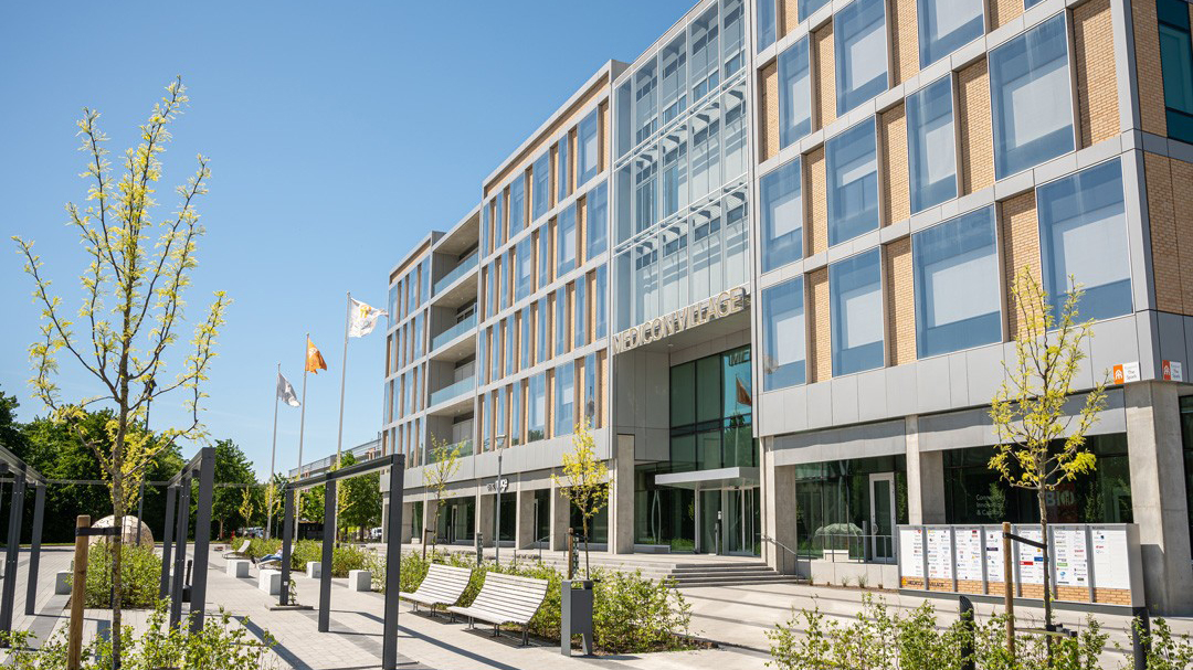 Framsidan av kontorsbyggnaden The Spark på Medicon Village med glasfasad, stora fönster och entré. Framför byggnaden syns en gångväg, träd, bänkar och flaggstänger i soligt väder.