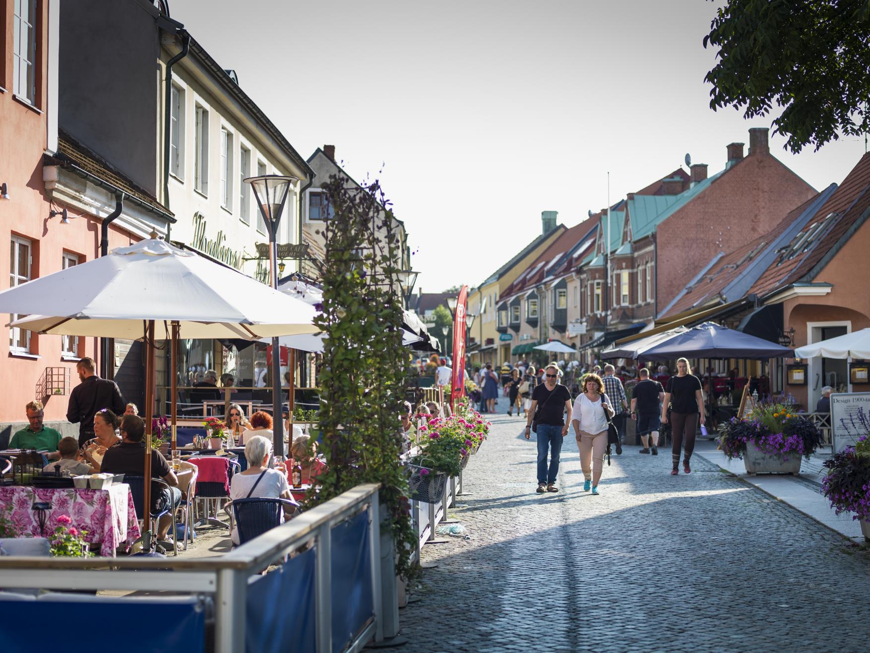 Kullerstensgata i europeisk stad med uteserveringar, människor som äter under parasoll och fotgängare som promenerar förbi färgglada byggnader prydda med blommor