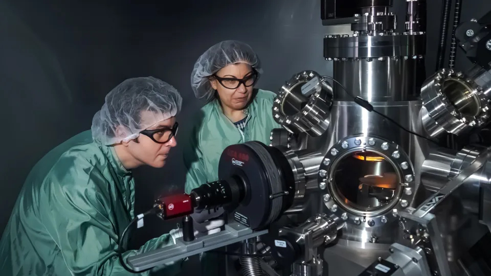 Two people with protective goggles, hairnets and labcoats are operating a machine. Photo.