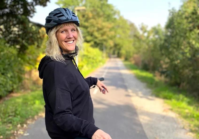 Foto av leende kvinna på cykel som står på en cykelväg och tittar bakåt in i kameran. Foto: Åsa Hansdotter.