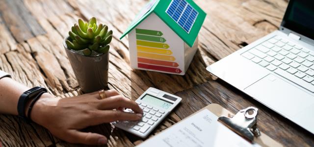 Person sitter med räkningar och miniräknare. Bredvid sig finns en liten modul av ett hus med staplar av energiklasser.