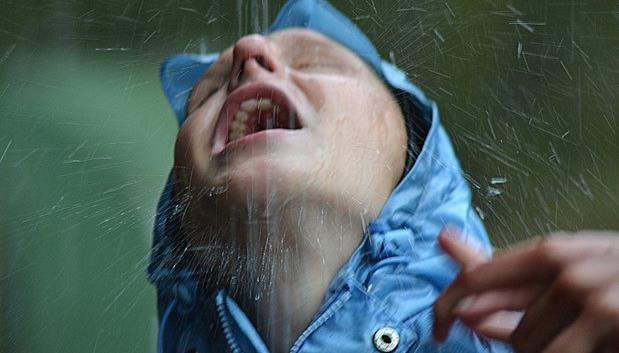 Ung mann med øynene lukket, står ute i regnet som renner over hans ansikt mens han er i taktil kontakt med en annen.