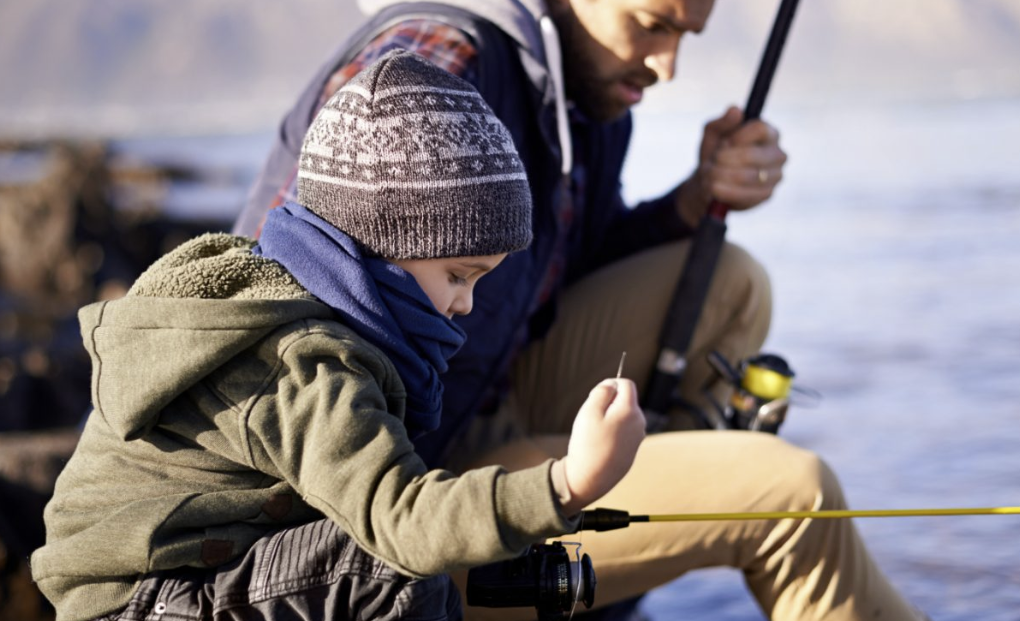 Adult man and young boy sitting outside by water fishing.