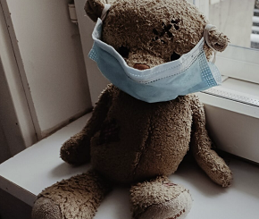 A teddy bear wearing a face mask sitting by a window.