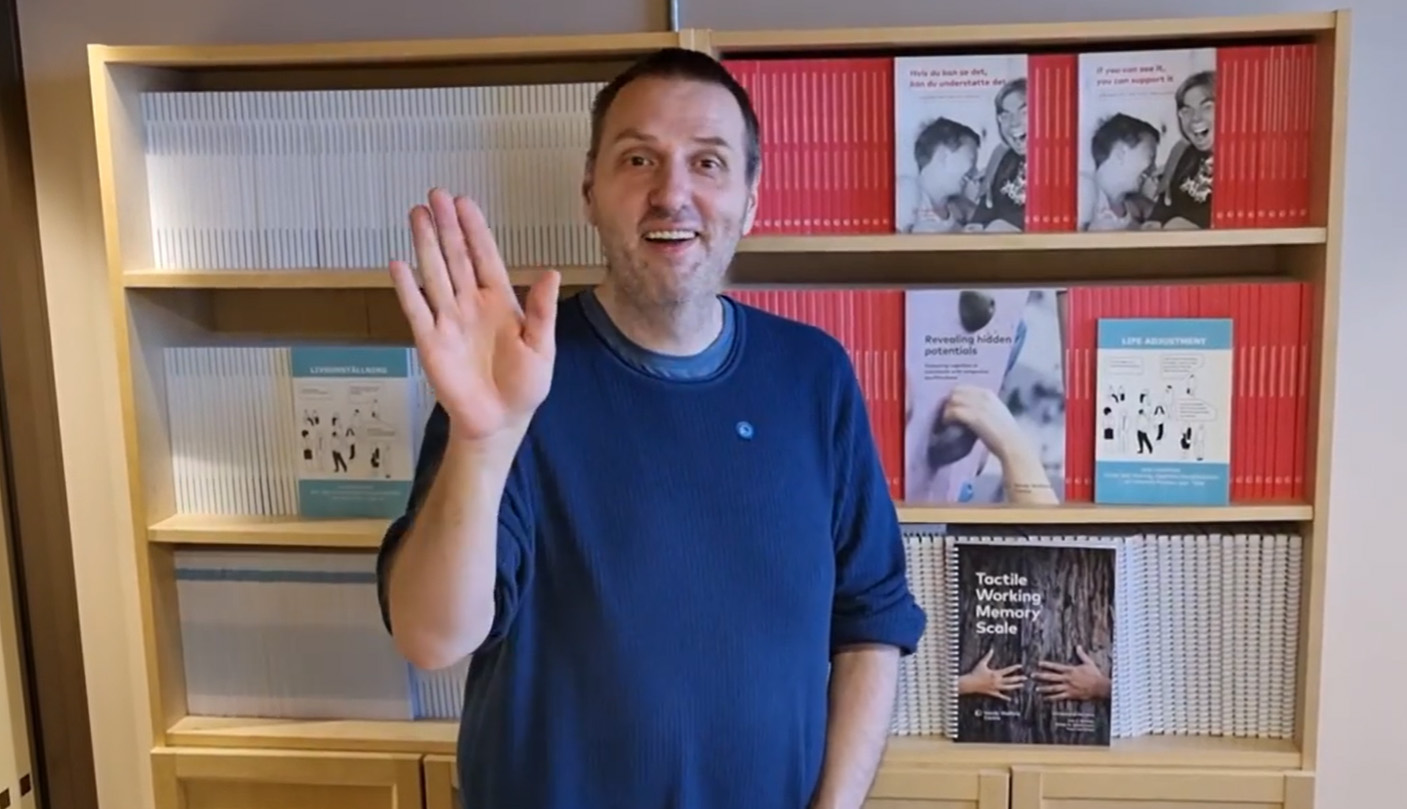 Bild på Göran Forsgren som står framför en bokhylla och vinkar med en hand. 