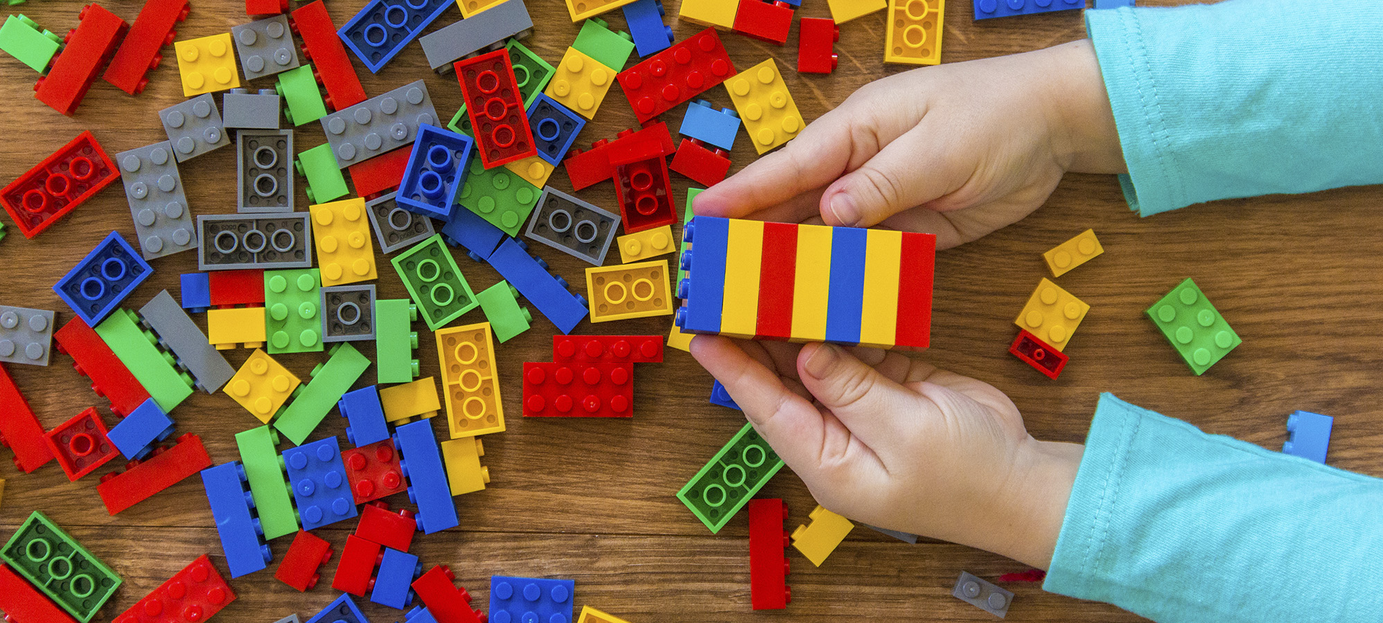  Et par hender holder en stabel Lego klosser over et bord fylt med Lego i forskjellige farger.