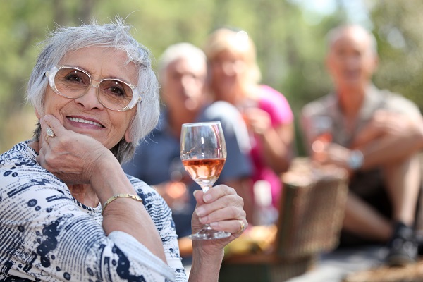 Äldre kvinna höjer vinglas mot kameran, vänner i bakgrunden på en picknickfilt