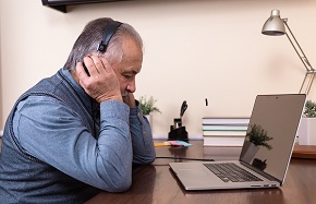 Äldre man sitter vid ett skrivbord och lyssnar på hörlurar vid sin dator.