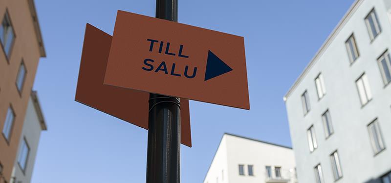 Till salu-skylt mot blå himmel och flerfamiljshus i bakgrunden.