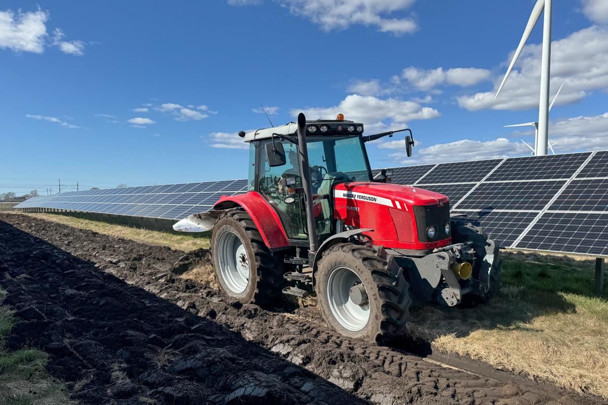 Traktor med plog som plöjer rader mellan två solcellsrader på en åker.