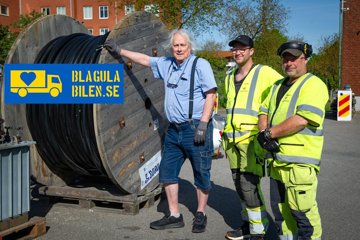 Tre män, två med gula skyddskläder, står uppställda bredvid en stor kabeltrumma för fotogarfering.