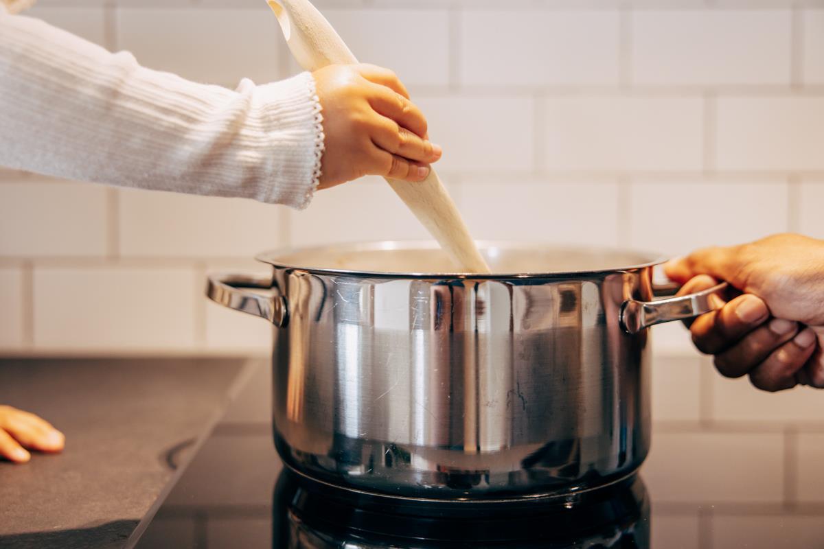 En barnhand rör med träslev i en gryta på en spis och vuxenhand håller i grytan också.