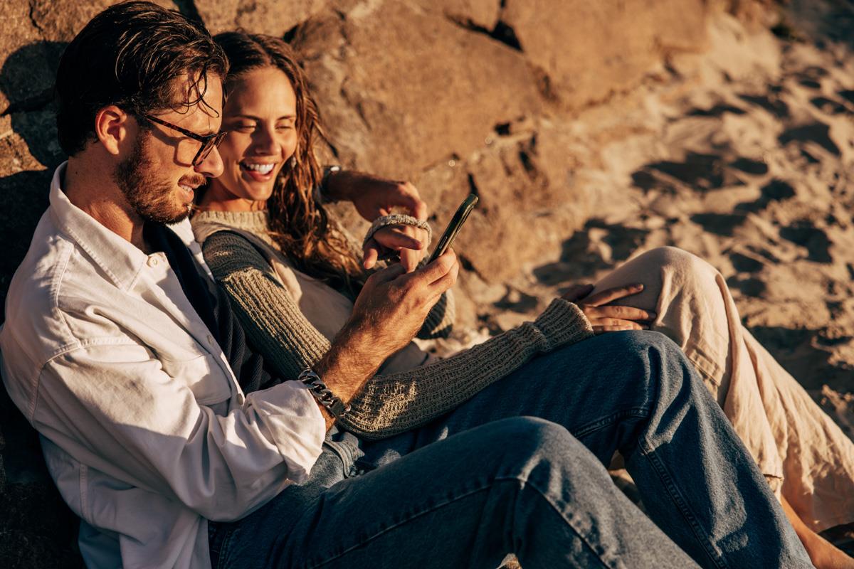 Man och kvinna sitter på en strand litande mot stenmur och tittar gemensamt i en mobil.