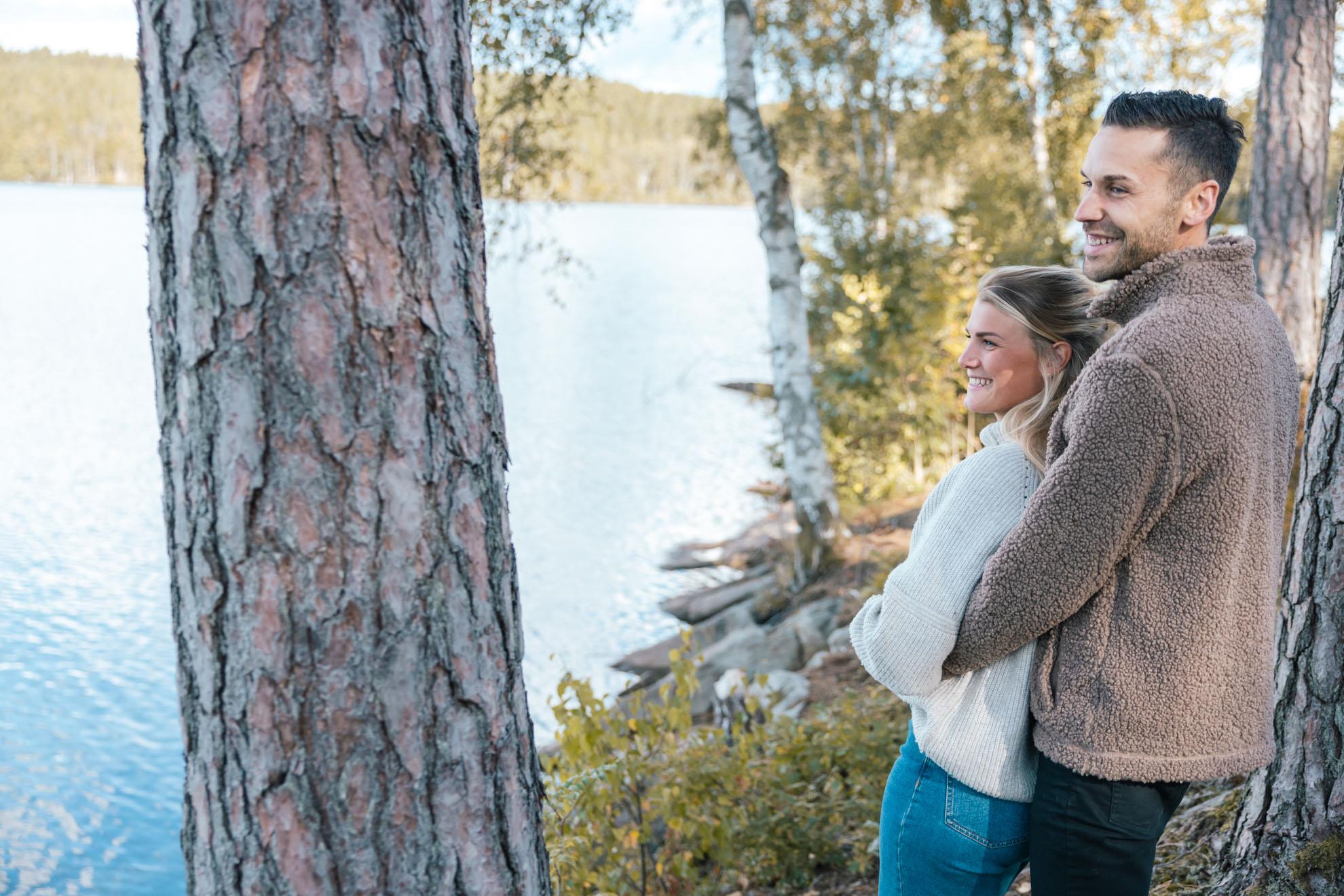 Två vuxna står och håller om varandra en sjö bredvid några tallar.